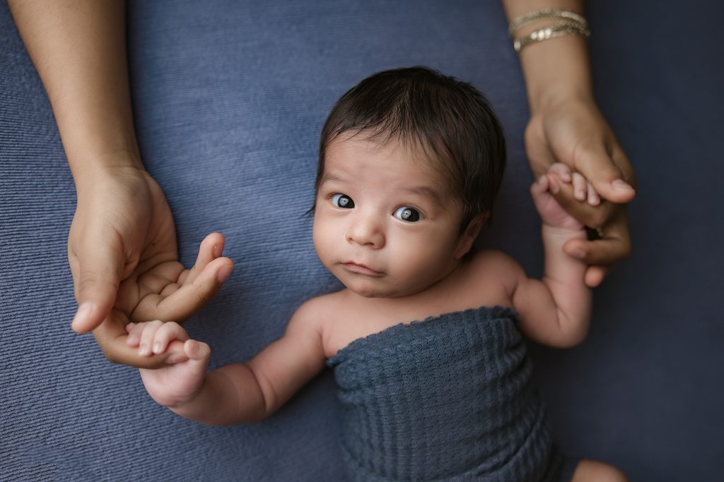 newborn-baby-boy-wrapped-in-blue-fabric-holding-moms-hands-during-newborn-photography-session-in-winston-salem-photography-studio