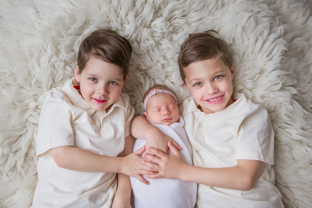 two-big-brothers-holding-newborn-baby-sister-laying-on-white-fuzzy-blanket-during-newborn-photo-session-greensboro-newborn-photo-studio
