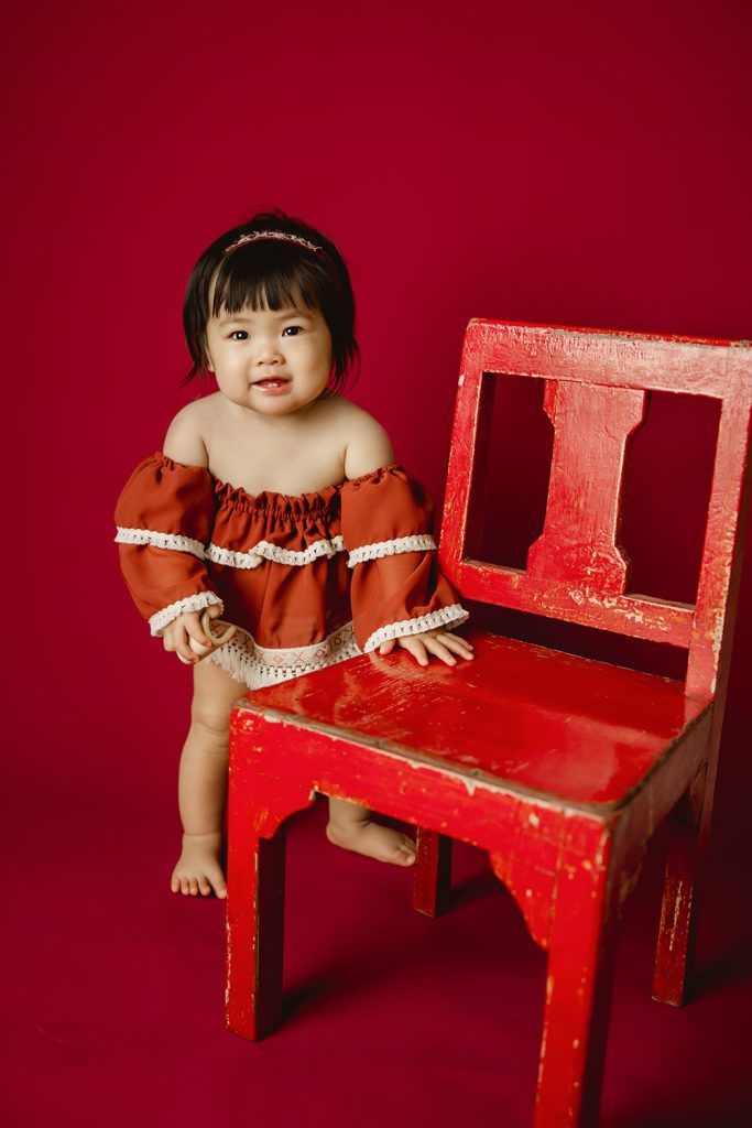 cute-baby-girl-in-red-outfit-standing-next-to-red-chair-in-red-background-smiling-during-greensboro-baby-photoshoot