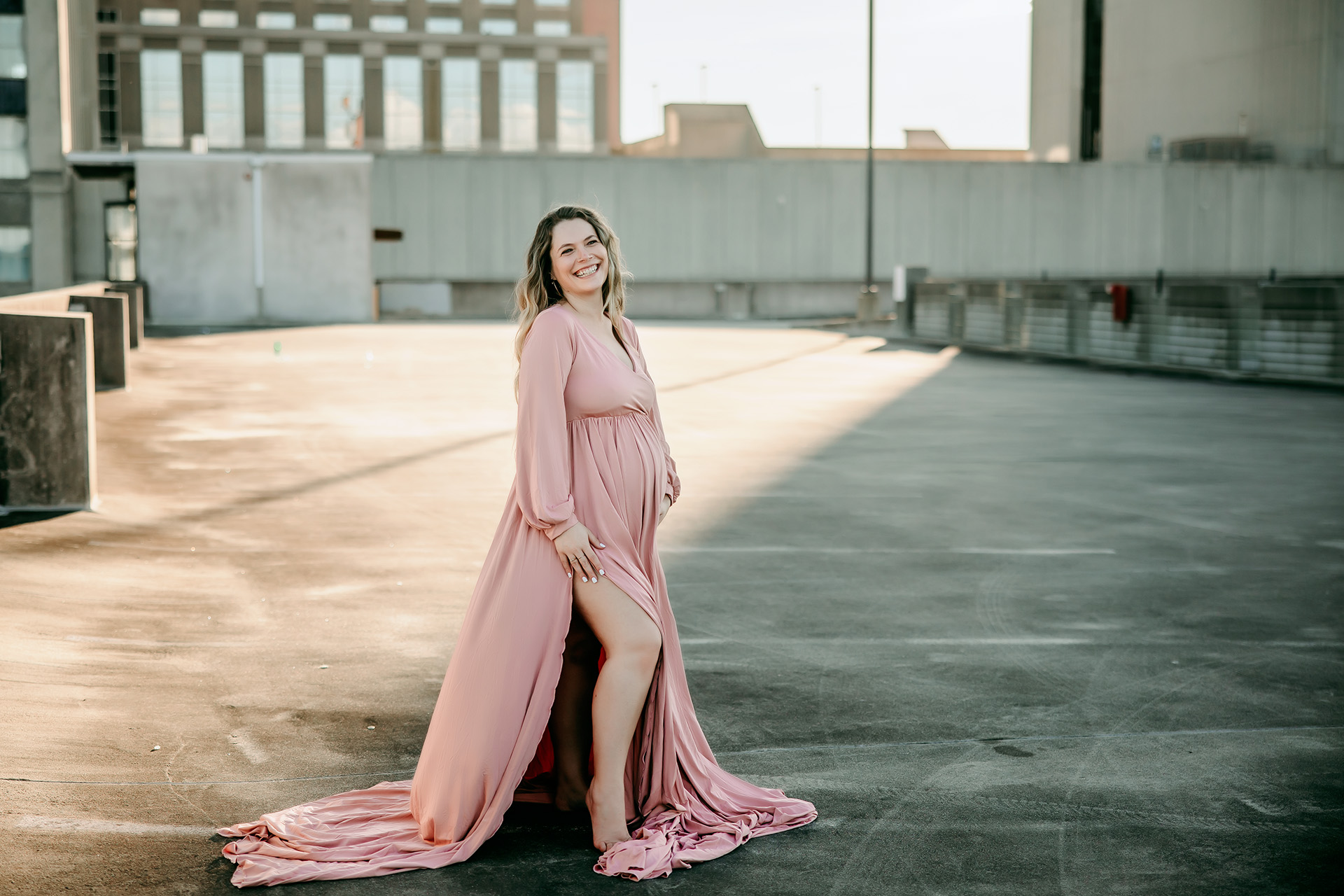 golden-hour-maternity-photoshoot-mama-smiling-in-pink-flowy-maternity-dress-roof-top-parking-lot-winston-salem