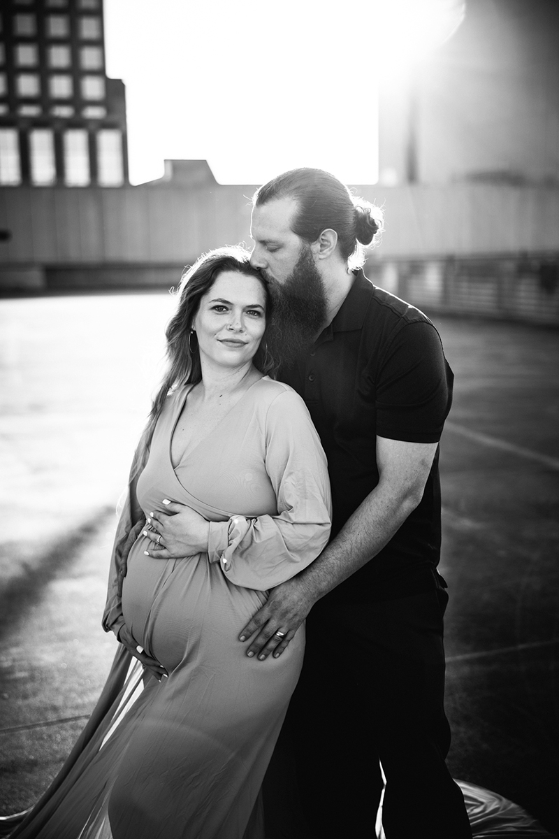 man-kissing-woman-during-golden-hour-maternity-photo-session-in-pink-flowy-maternity-dress-roof-top-parking-lot-winston-salem-downtown