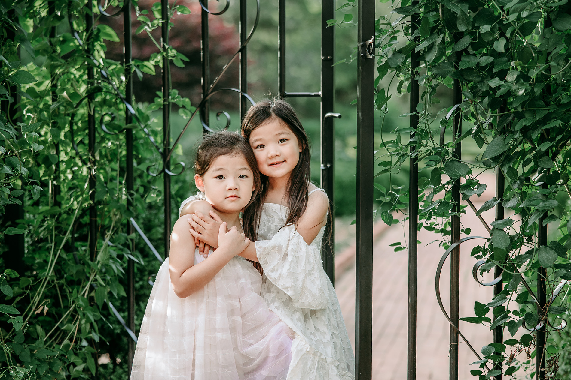 Two-pretty-asian-sister-in-white-dresses-hugging-each-other-in-front-of-iron-gate-in-Tanglewood-garden-winston-salem-family-photography