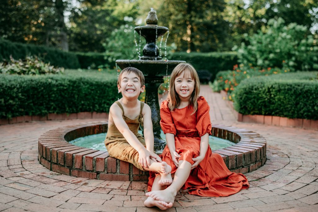 brother and sister sitting in front of the fountain smiling during family photo session in Tanglewood botanic garden with Greensboro family photographer