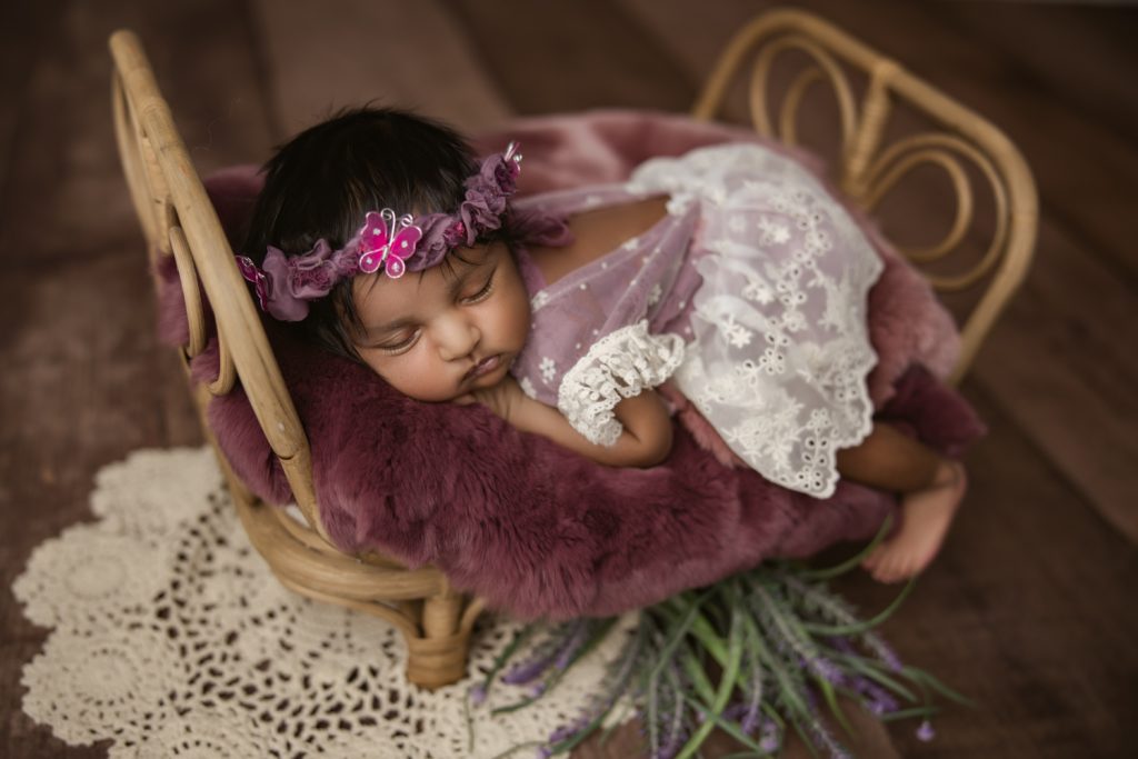 newborn baby girl in berry dress and bonnet sleeping on a bamboo bed during newborn photo session Charlotte