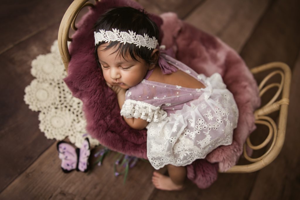 newborn baby girl in berry dress and bonnet sleeping on a bamboo bed during newborn photo session Charlotte