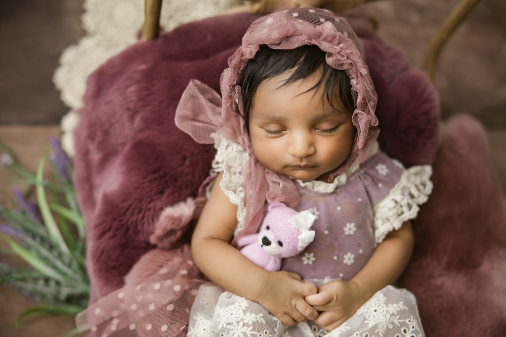 newborn baby girl in berry dress and bonnet sleeping on a bamboo bed during newborn photo session greensboro