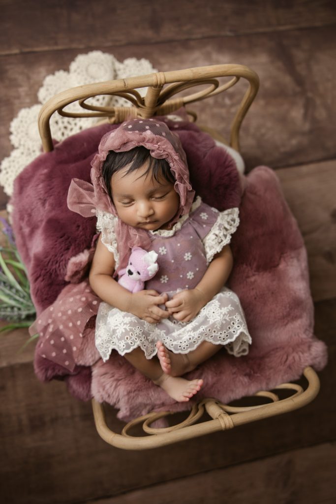 newborn baby girl in berry dress and bonnet sleeping on a bamboo bed during newborn photo session greensboro