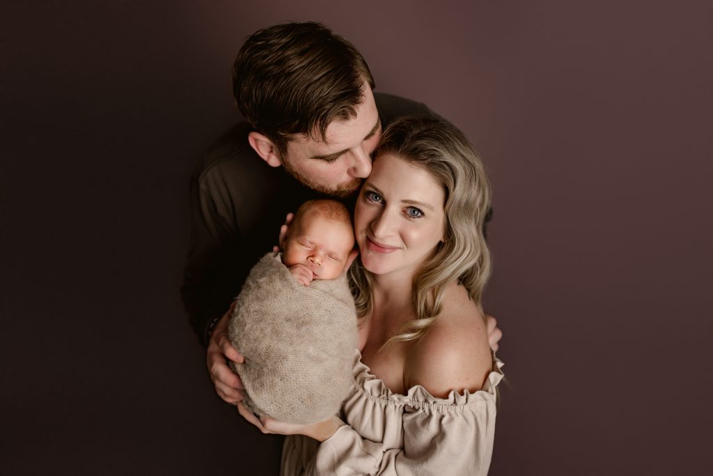 dad kissing mom while mom holding their newborn baby boy for family photo during Winston Salem newborn photography session