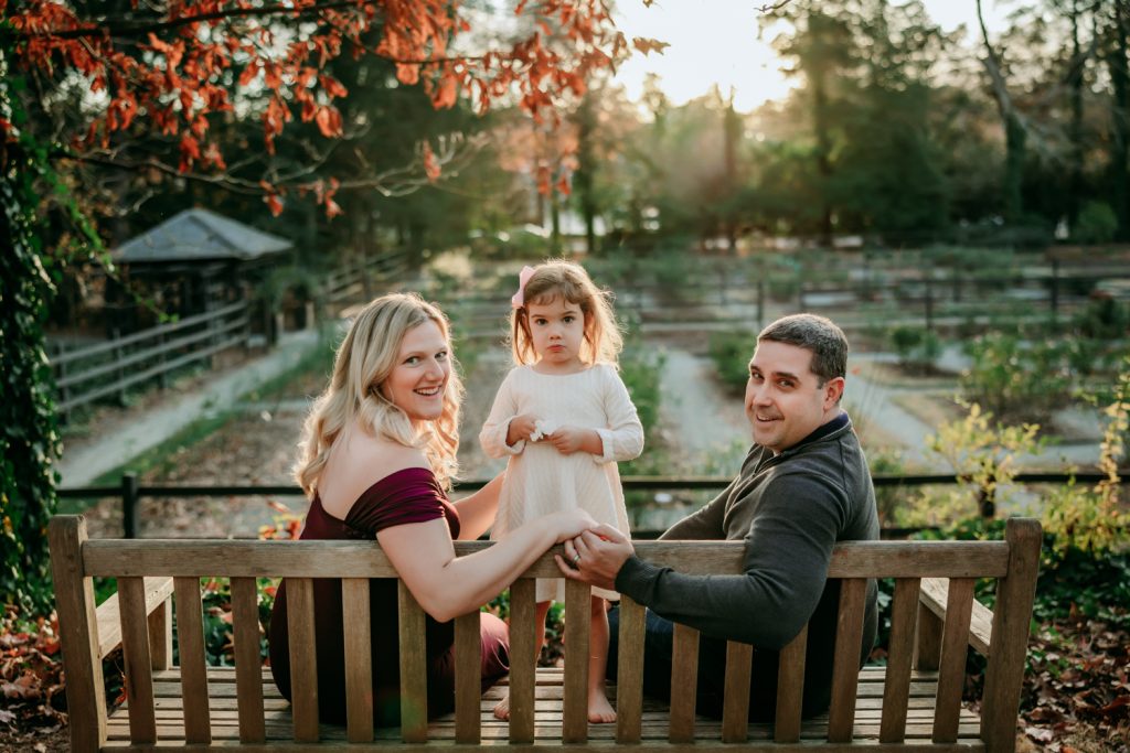mom and dad with little girl looking back while sitting on the bench during sunset family photography in Reynolda garden with Winston salem photographer