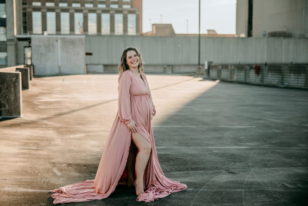 golden hour maternity photo session with expectant mother in flowy pink dress at the rooftop parking lot in downtown Winston Salem with greensboro maternity photographer