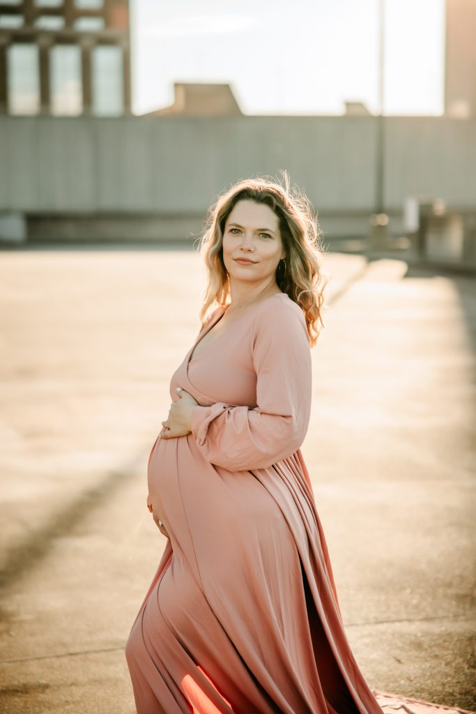 golden hour maternity photo session with expectant mother in flowy pink dress at the rooftop parking lot in downtown Winston Salem with greensboro maternity photographer