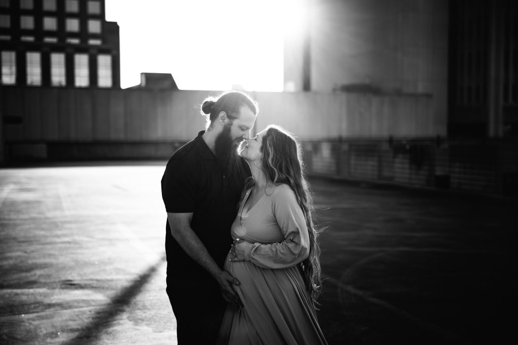 Black and white maternity photo of expectant mother with her husband kissing each other at the rooftop parking lot in downtown Winston Salem with greensboro maternity photographer