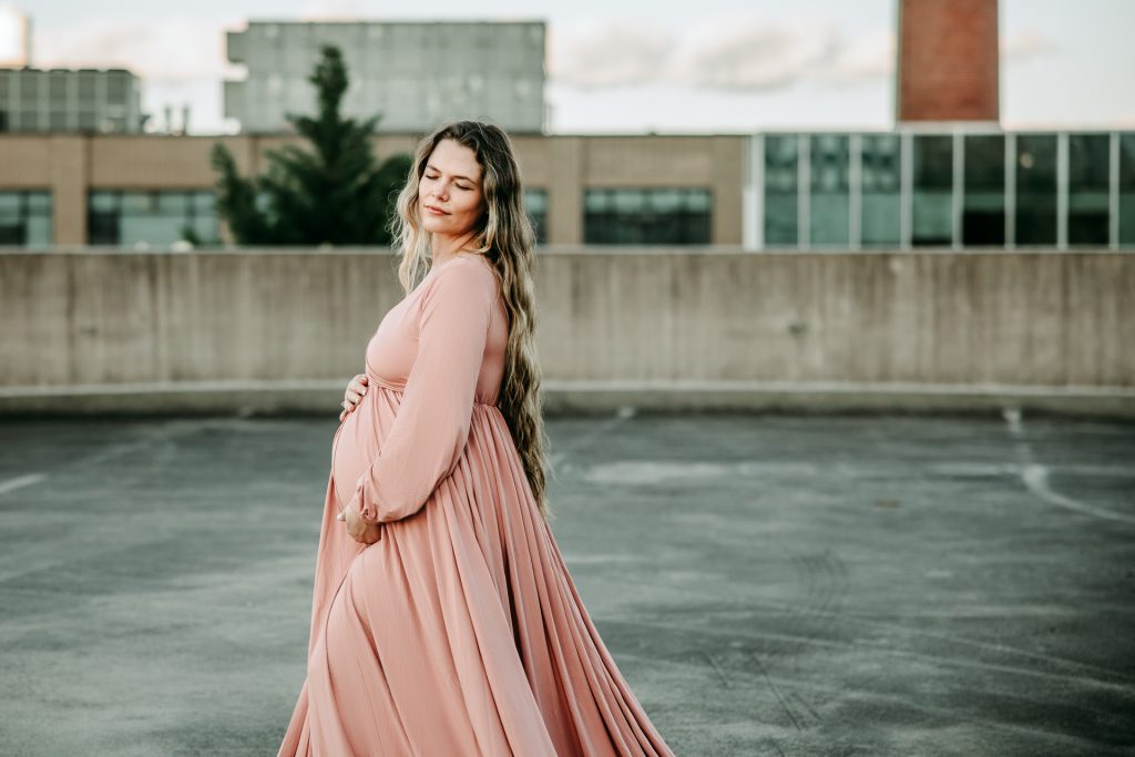golden hour maternity photo session with expectant mother in flowy pink dress at the rooftop parking lot in downtown Winston Salem with greensboro maternity photographer