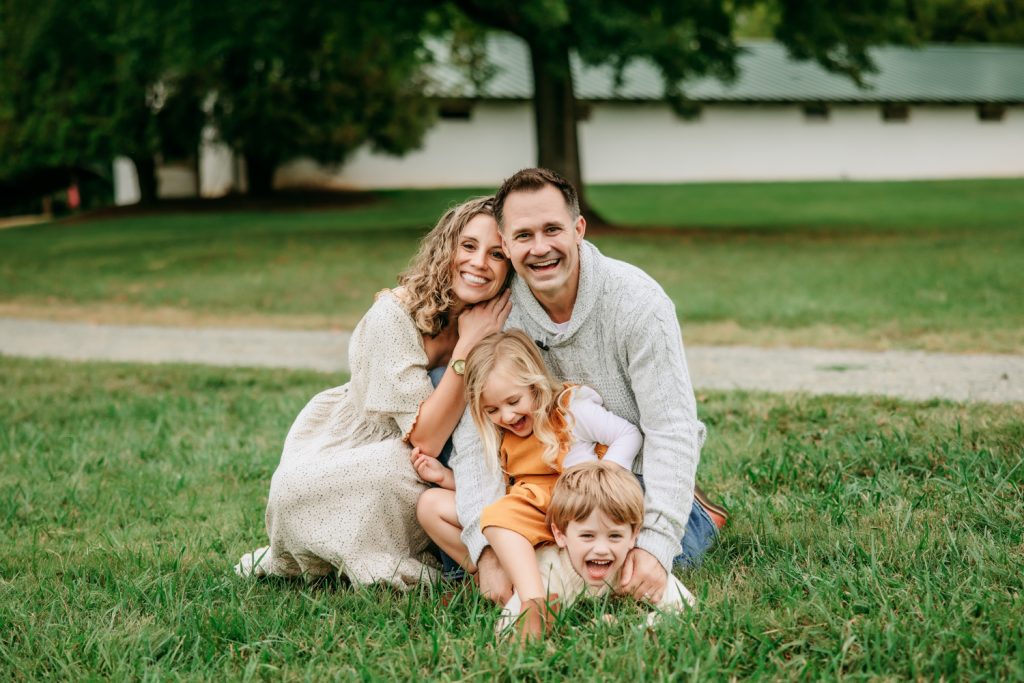 mom and dad sitting down on the grass with daughter and son smiling in front of a horse barn during family photo session at Tanglewood Park with greensboro family photographer