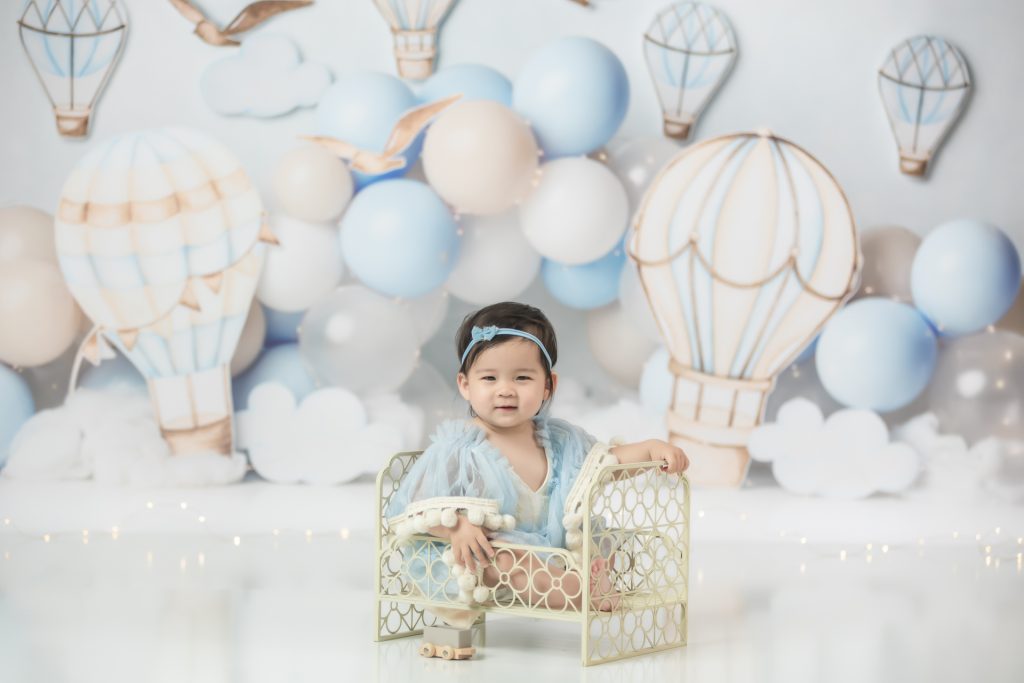 baby girl in blue dress sitting in white iron bed during hot air balloon theme first birthday session Winston Salem baby photographer
