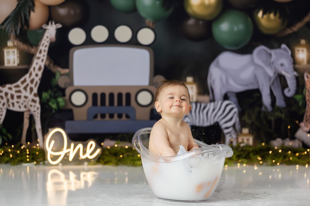baby boy in milk bath sucking his fingers during first birthday cake smash photoshoot with greensboro baby photographer