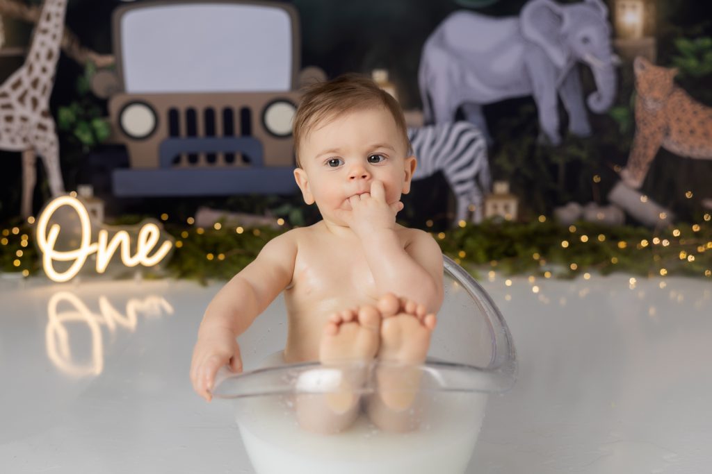 baby boy in milk bath sucking his fingers during first birthday cake smash photoshoot with greensboro baby photographer