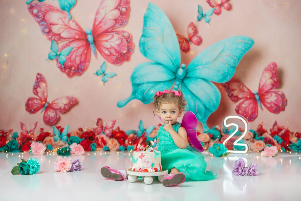cute baby girl in teal dress and fuchsia wings during second birthday cake smash session in butterfly themed with greensboro baby photographer