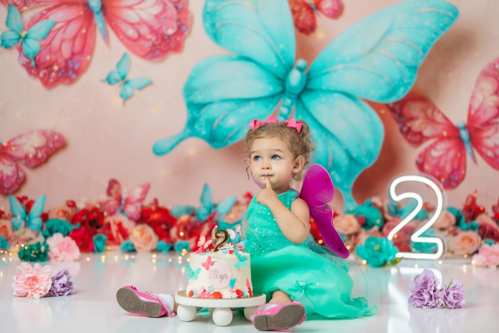 cute baby girl in teal dress and fuchsia wings during second birthday cake smash session in butterfly themed with greensboro baby photographer