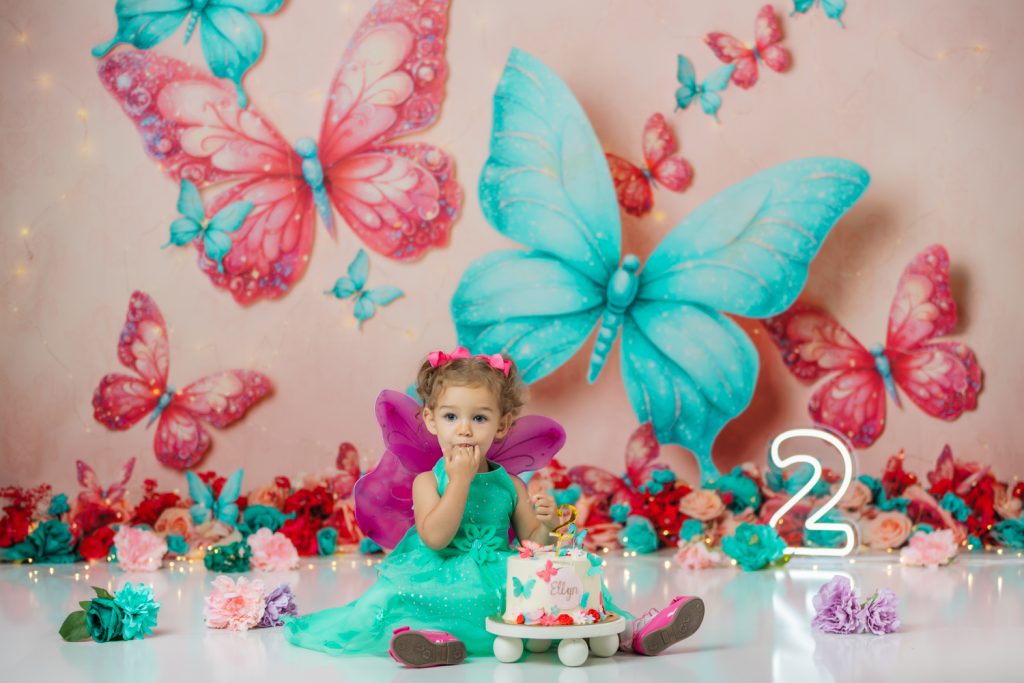 cute baby girl in teal dress and fuchsia wings during second birthday cake smash session in butterfly themed with greensboro baby photographer