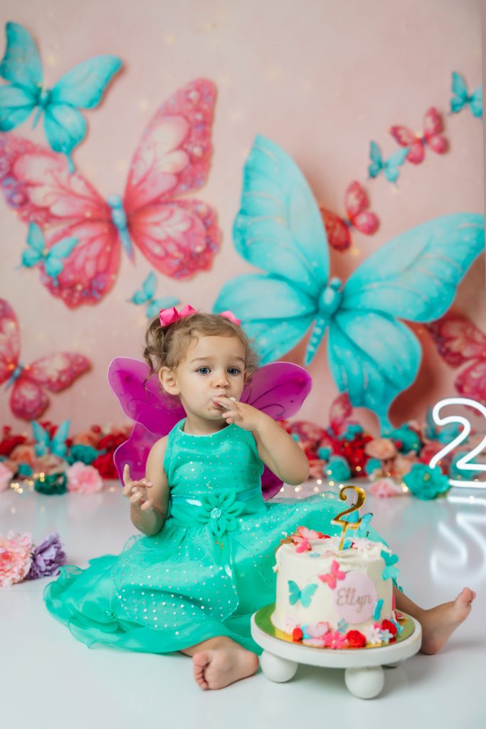 cute baby girl in teal dress and fuchsia wings during second birthday cake smash session in butterfly themed with greensboro baby photographer