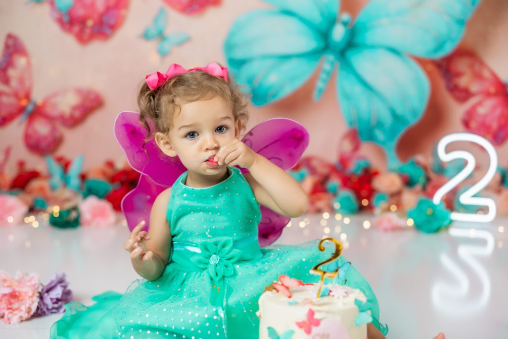 cute baby girl in teal dress and fuchsia wings during second birthday cake smash session in butterfly themed with greensboro baby photographer