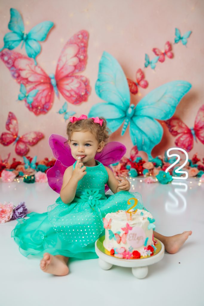 cute baby girl in teal dress and fuchsia wings during second birthday cake smash session in butterfly themed with greensboro baby photographer