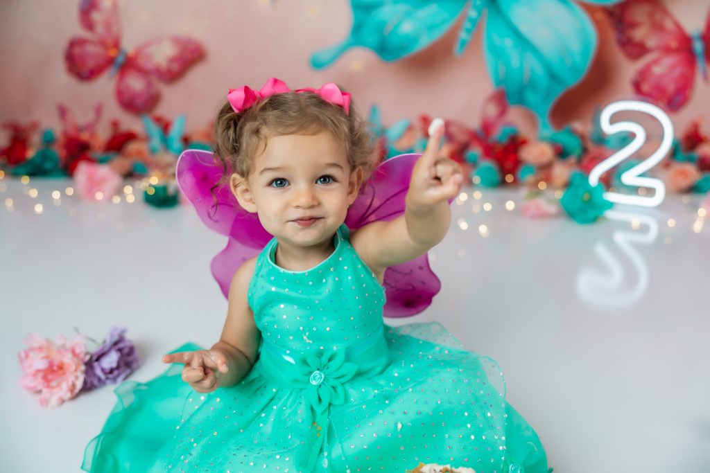 cute baby girl in teal dress and fuchsia wings during second birthday cake smash session in butterfly themed with greensboro baby photographer