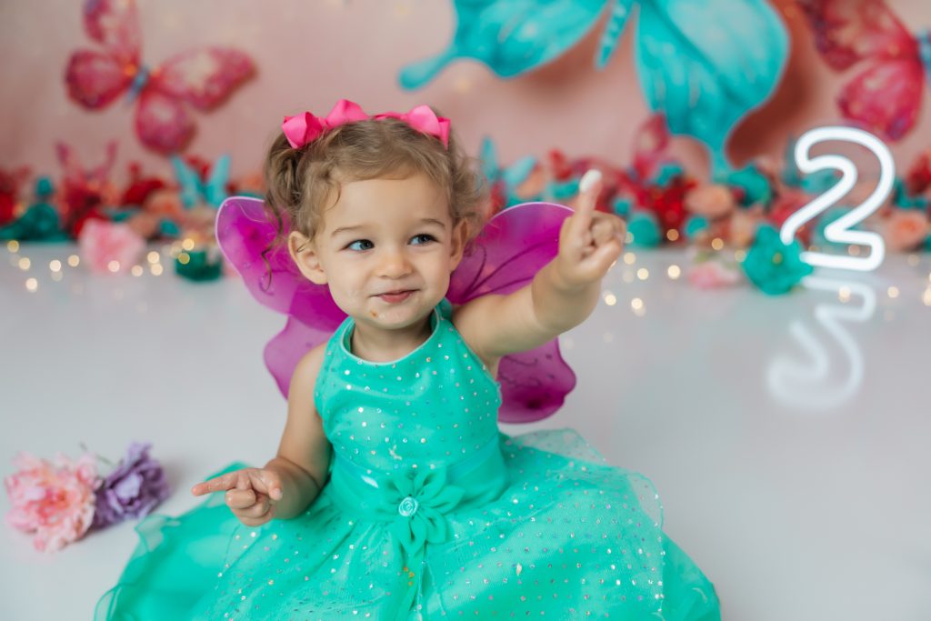 cute baby girl in teal dress and fuchsia wings during second birthday cake smash session in butterfly themed with greensboro baby photographer