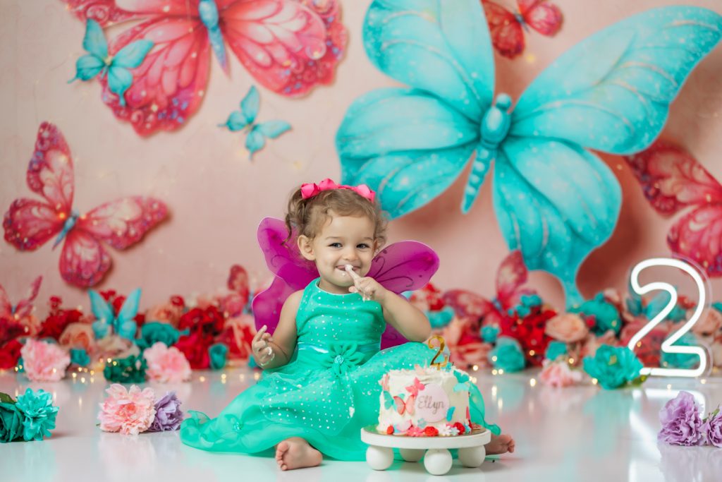 cute baby girl in teal dress and fuchsia wings during second birthday cake smash session in butterfly themed with greensboro baby photographer