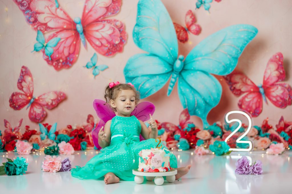 cute baby girl in teal dress and fuchsia wings during second birthday cake smash session in butterfly themed with greensboro baby photographer