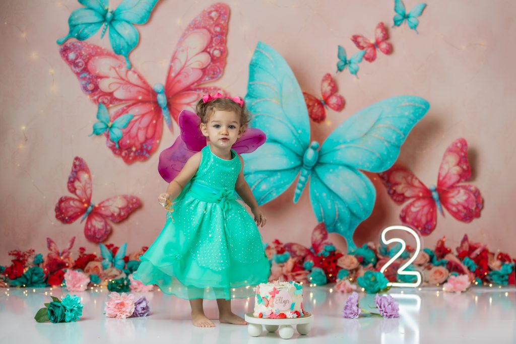 cute baby girl in teal dress and fuchsia wings during second birthday cake smash session in butterfly themed with greensboro baby photographer