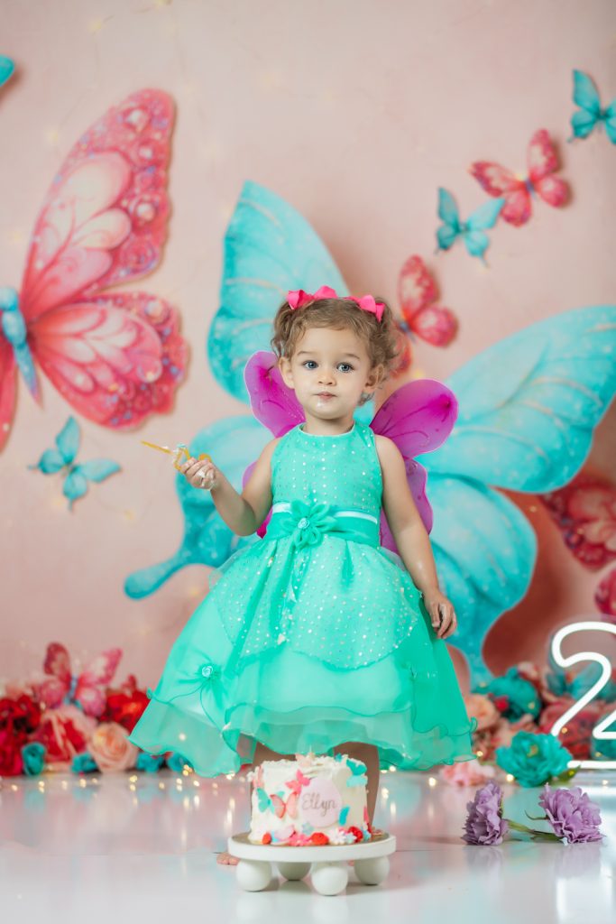 cute baby girl in teal dress and fuchsia wings during second birthday cake smash session in butterfly themed with greensboro baby photographer