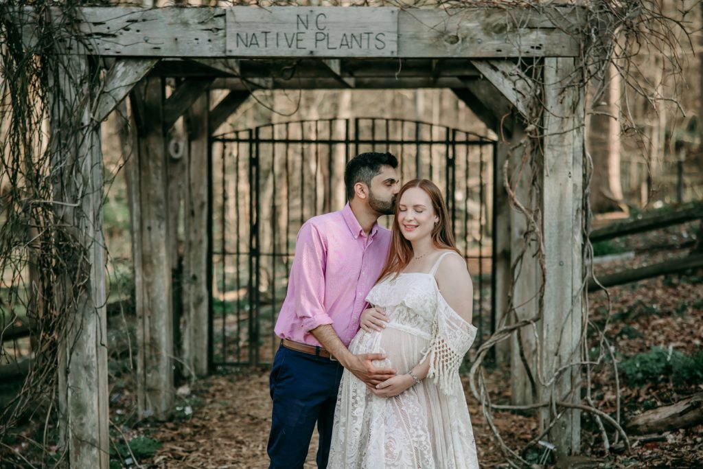 husband-kissing-his pregnant-wife-during-golden-hour-maternity-photo-session-in-tanglewood-park-arboretum-winston-salem-NC
