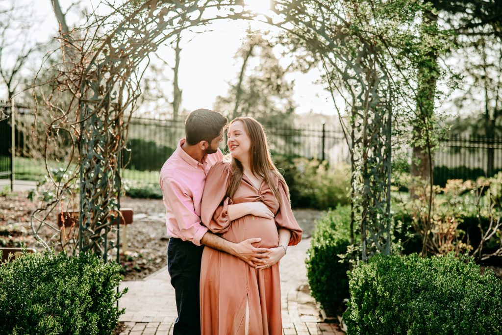 wife-and-husband-looking-at-each-other-with-smile-in-tanglewood-park-garden-during-maternity-photo-session-with-winston-salem-motherhood-photographer