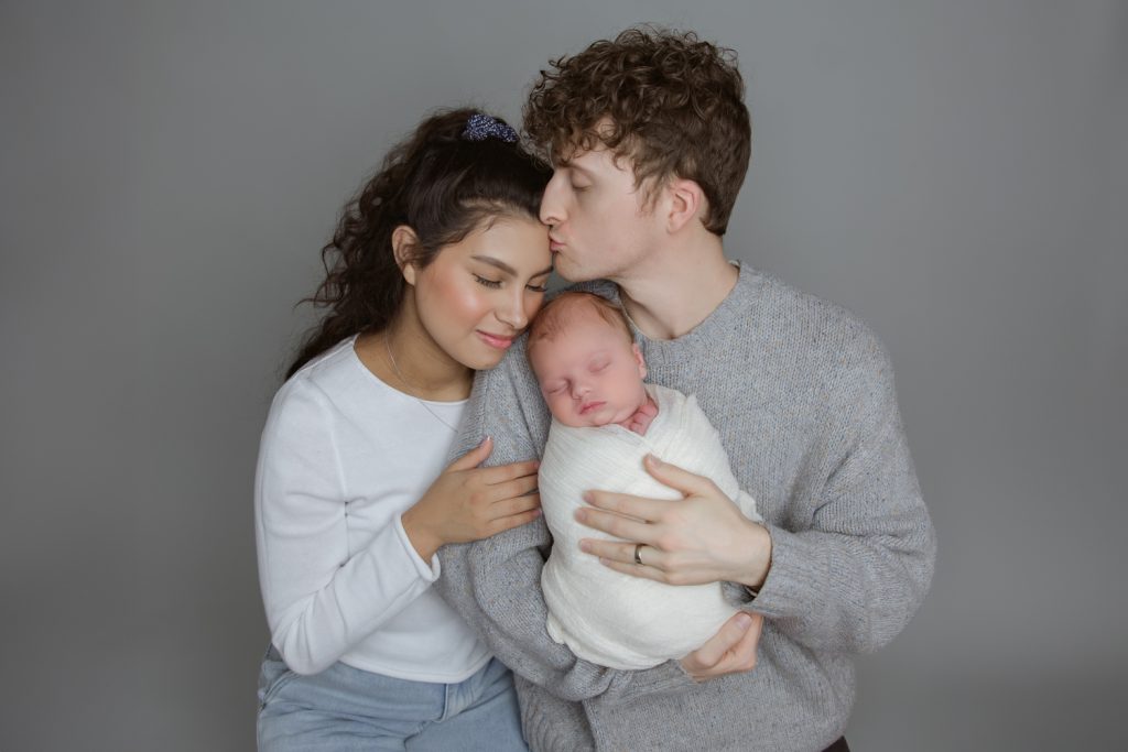 dad kissing mom on her forehead while holding newborn baby boy during family photo with Winston Salem newborn photographer