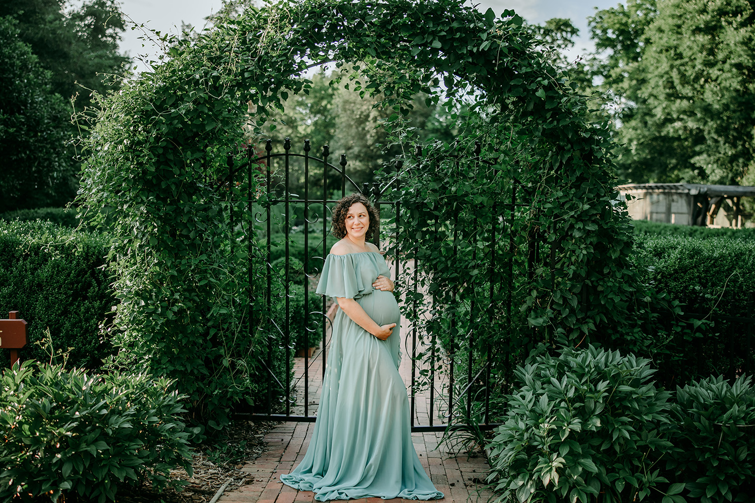 maternity mother dressed in teal gown in front of a beautiful iron gate at Tanglewood park Clemmons NC during maternity photo shoot with Winston Salem maternity photographer