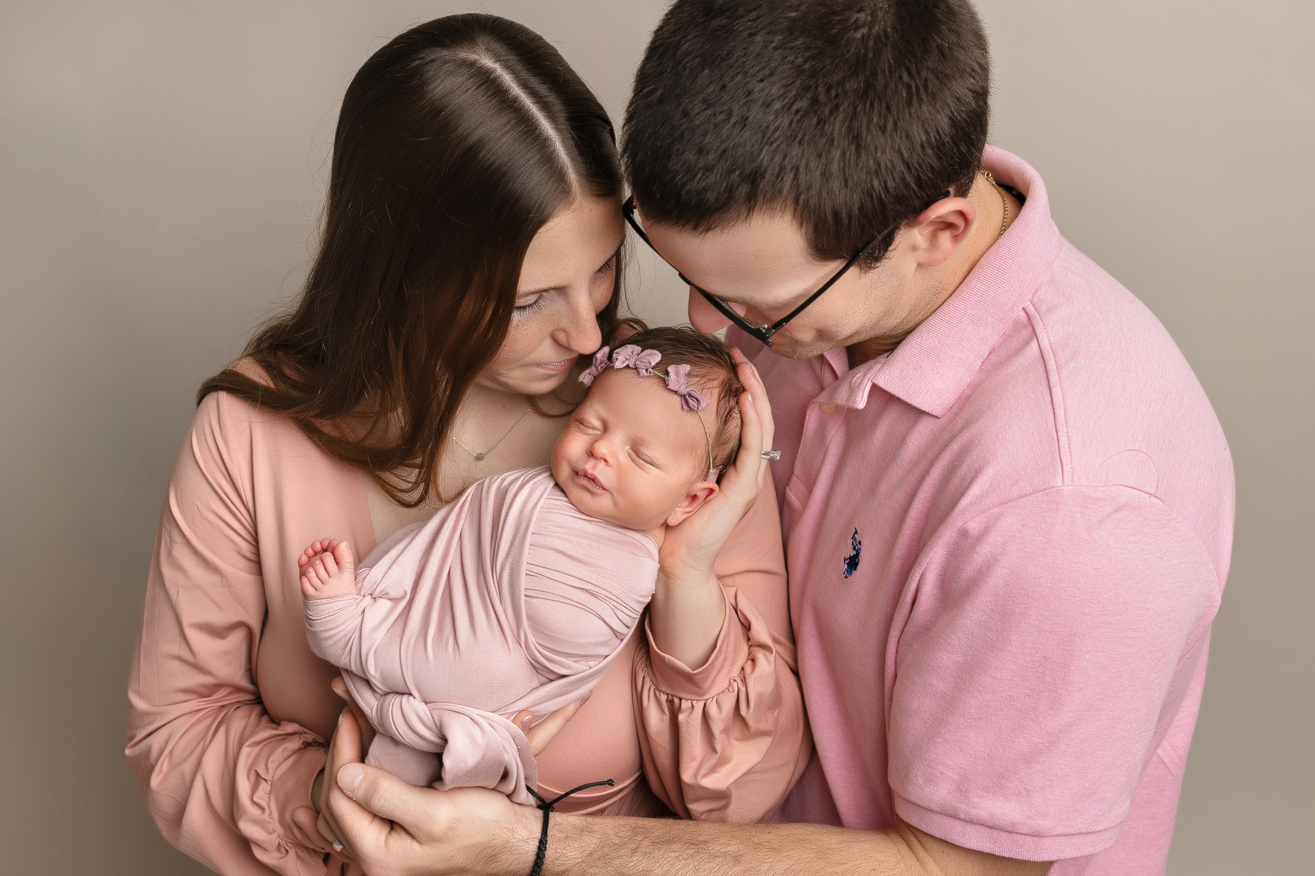 mom-and-dad-holding-newborn-baby-girl-all-dressed-in-pink-outfits-mom-and-dad-looking-down-on-baby-during-greensboro-newborn-photo-shoot