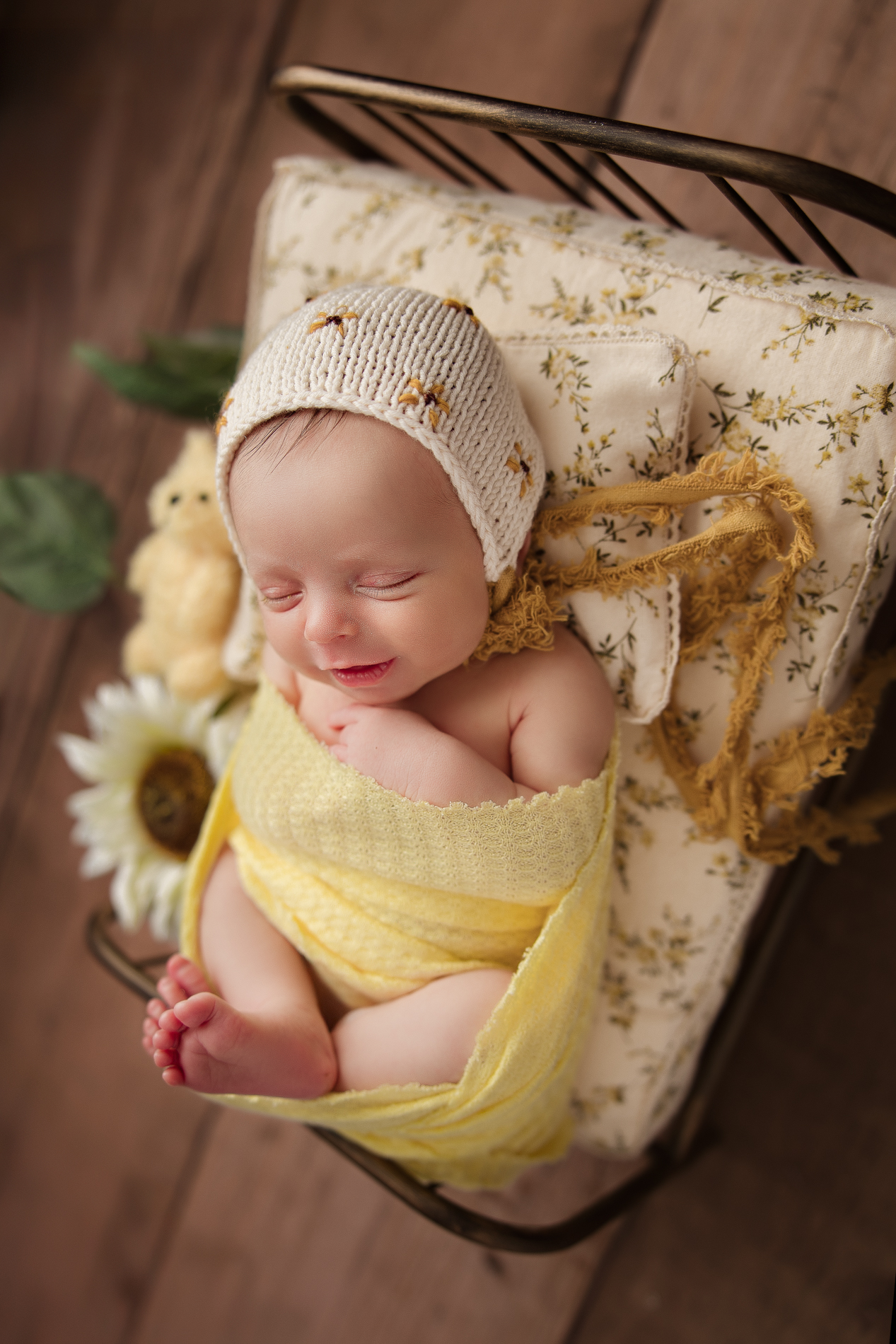 newborn-baby-smiling-in-sleep-swaddled-in-yellow-fabric-wearing-a-daisy-bonnet-laying-in-a-tiny-bed-with-sunflowers-during-newborn-baby-photography-session-greensboro-photographer