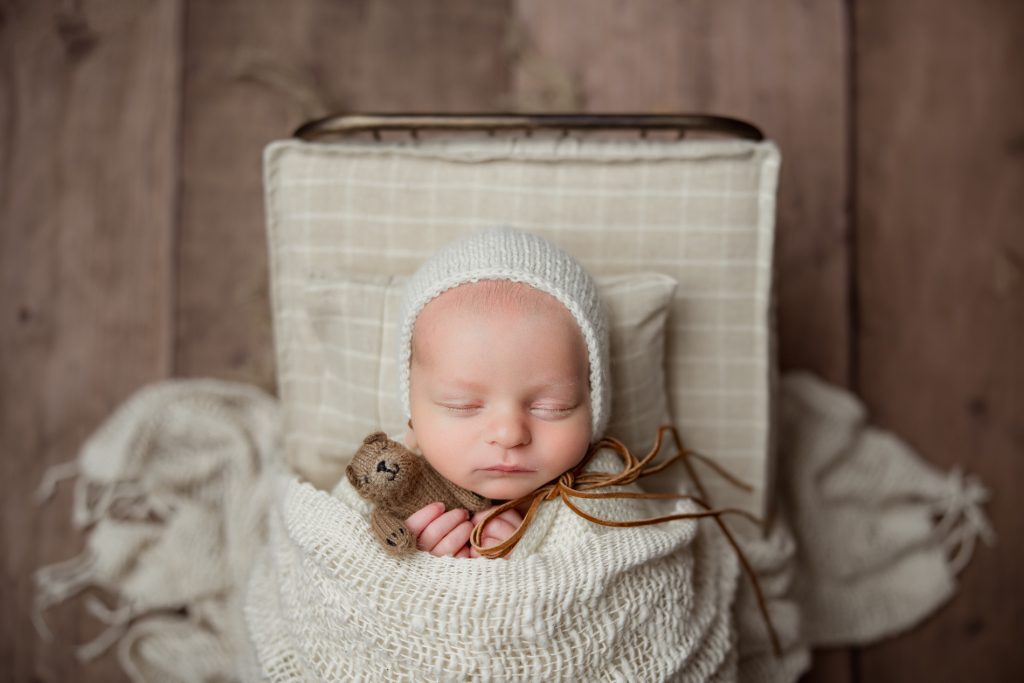 newborn-baby-boy-sleeping-in-a-white-baby-bed-tucked-in-holding-brown-teddy-bear-during-winston-salem-newborn-photography-session