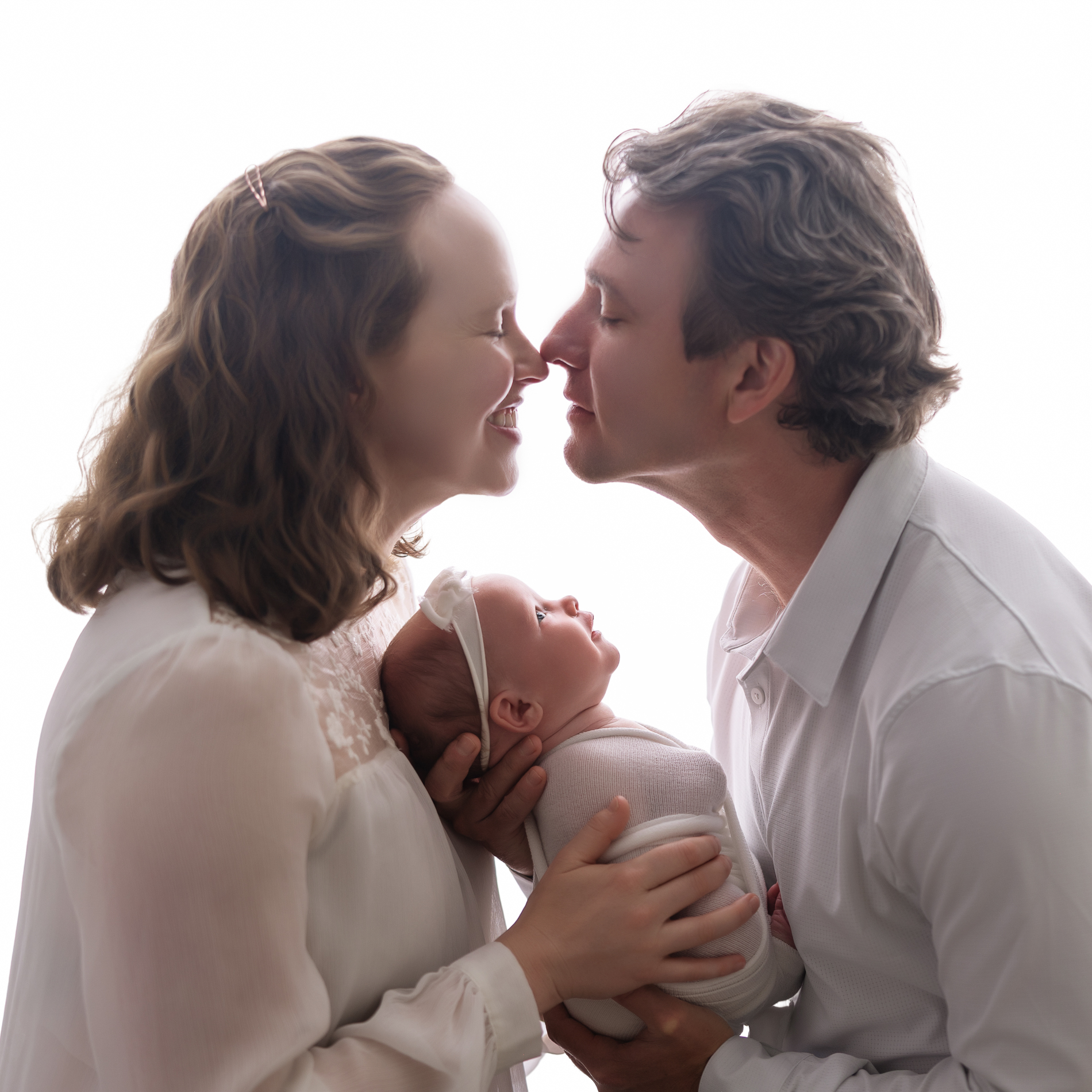 parents-kissing-during-newborn-photoshoot-while-holding-baby-girl-in-white-background-with-winston-salem-newborn-photographer
